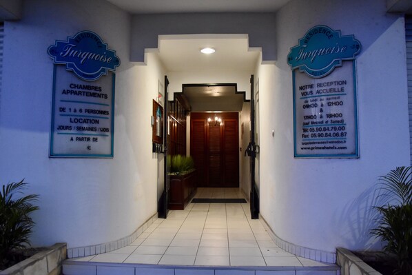 A hotel entrance with a reception desk, a staircase, and two signs indicating the hotel name and reception hours.