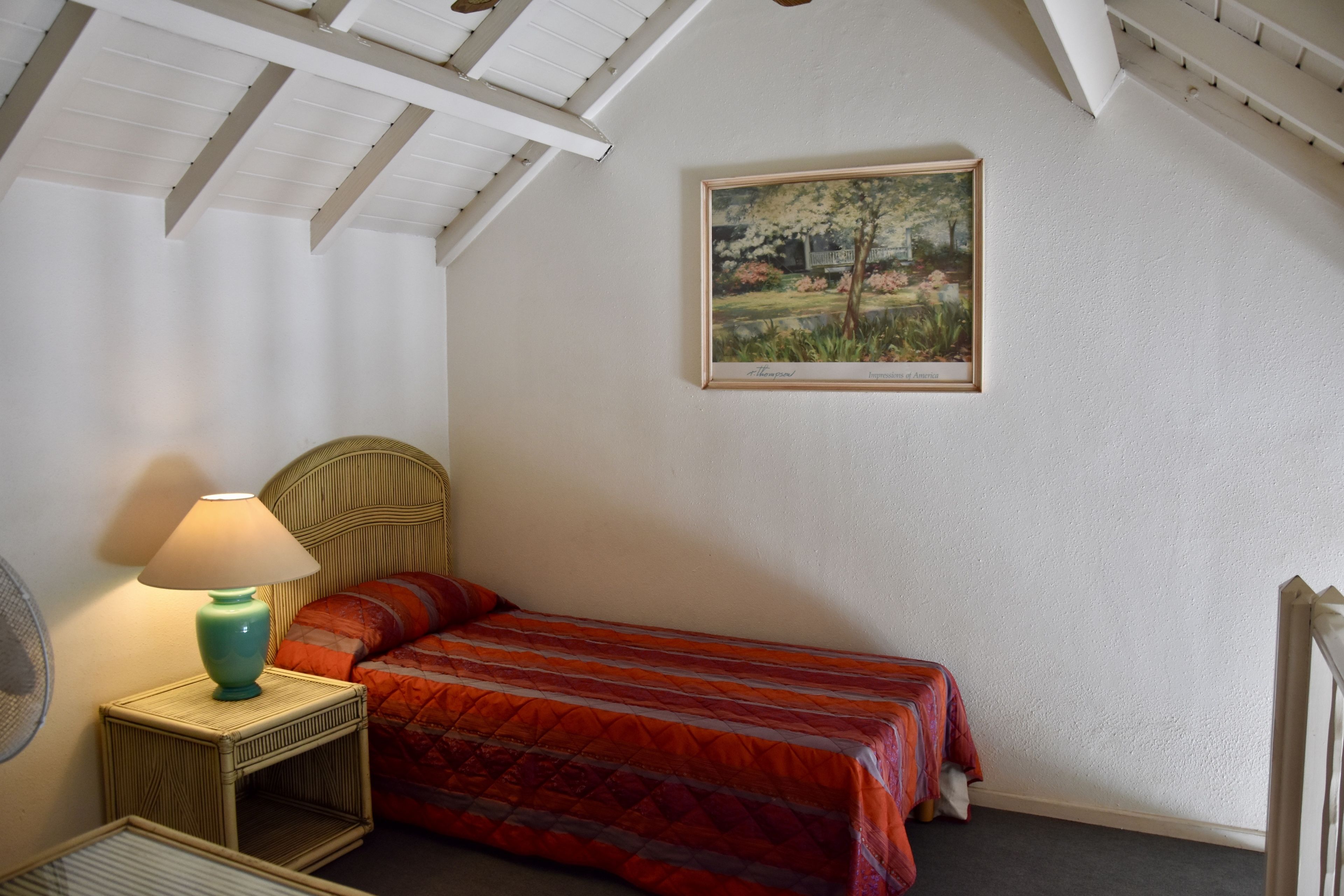 Une chambre avec un lit simple, une table de chevet avec une lampe, un tableau encadré au mur et un ventilateur de plafond.