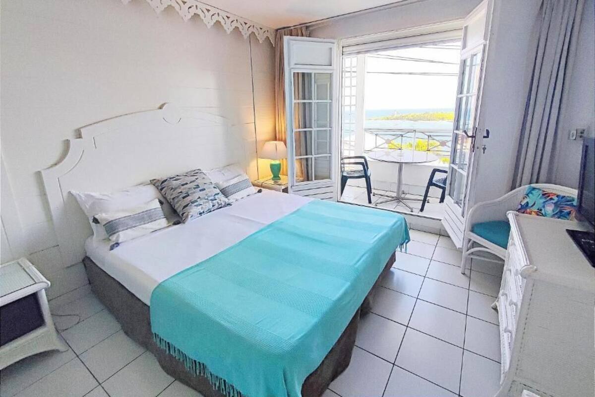 A bedroom with a bed, a bedside table, a chair, a small table with a lamp, and a balcony with a table and chairs.