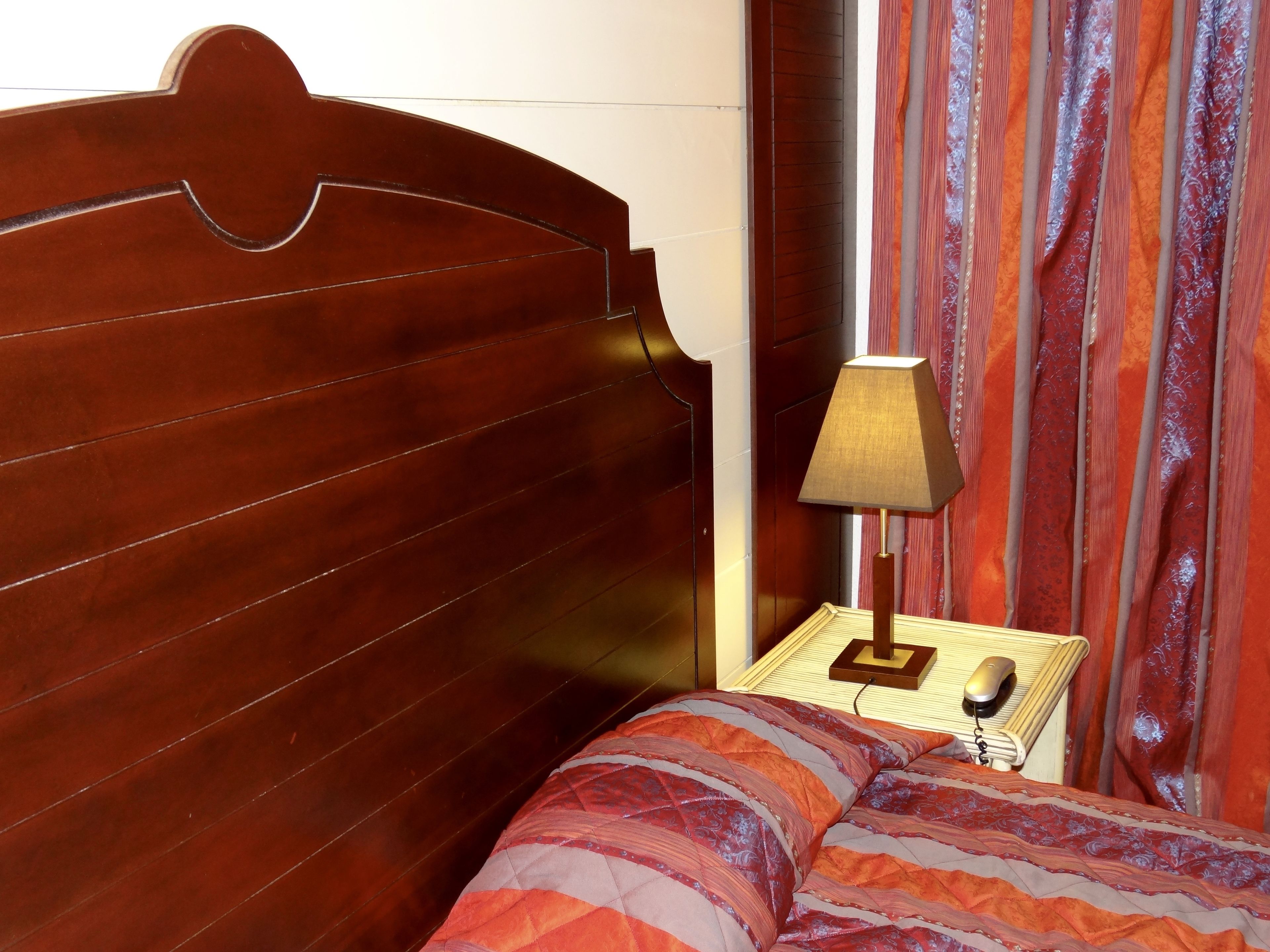 A wooden headboard, a bedside lamp, a patterned bedspread, and a nightstand with a phone.
