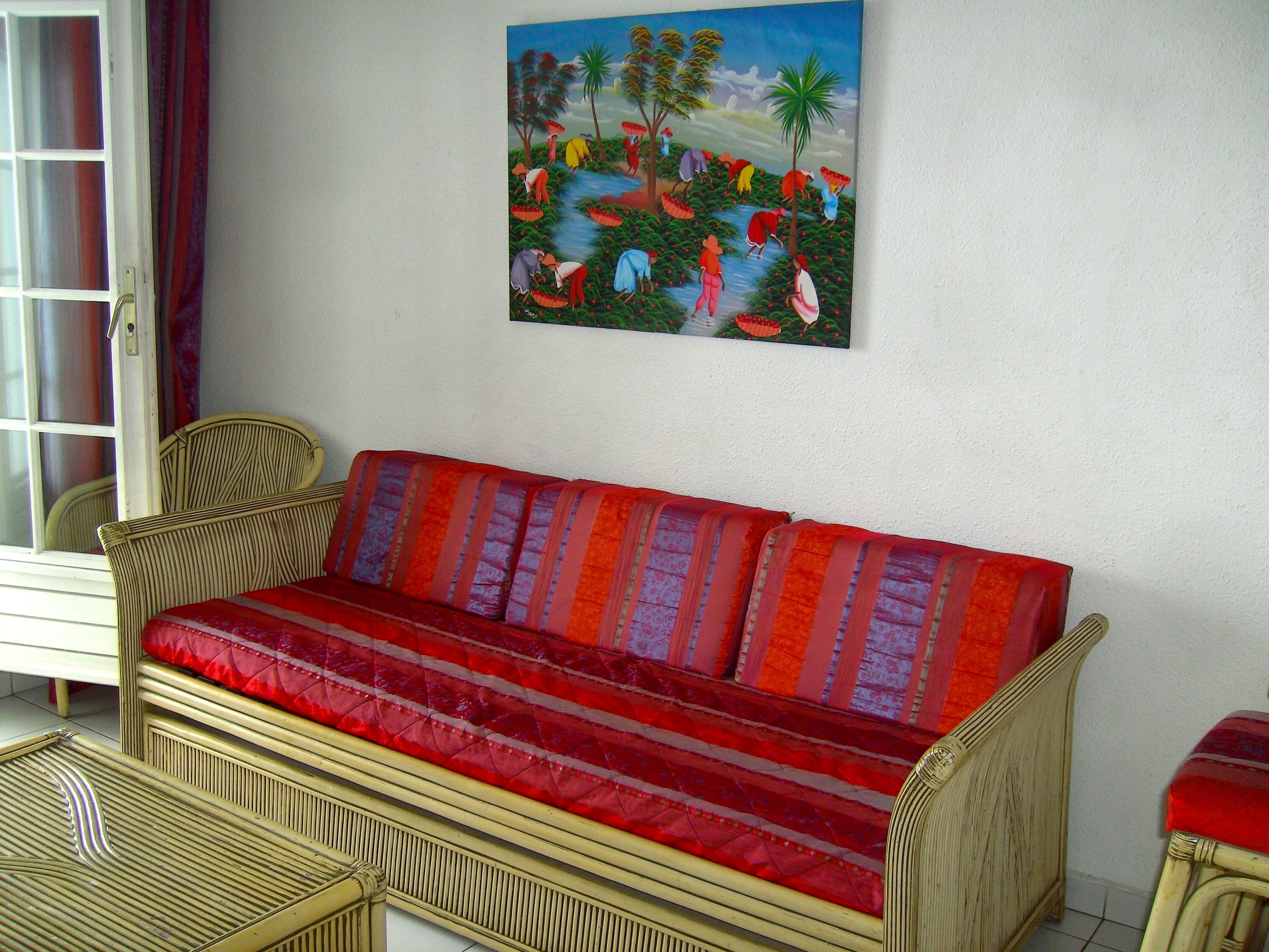 Une pièce avec un canapé en osier, un tableau représentant un paysage tropical et une fenêtre avec des rideaux.