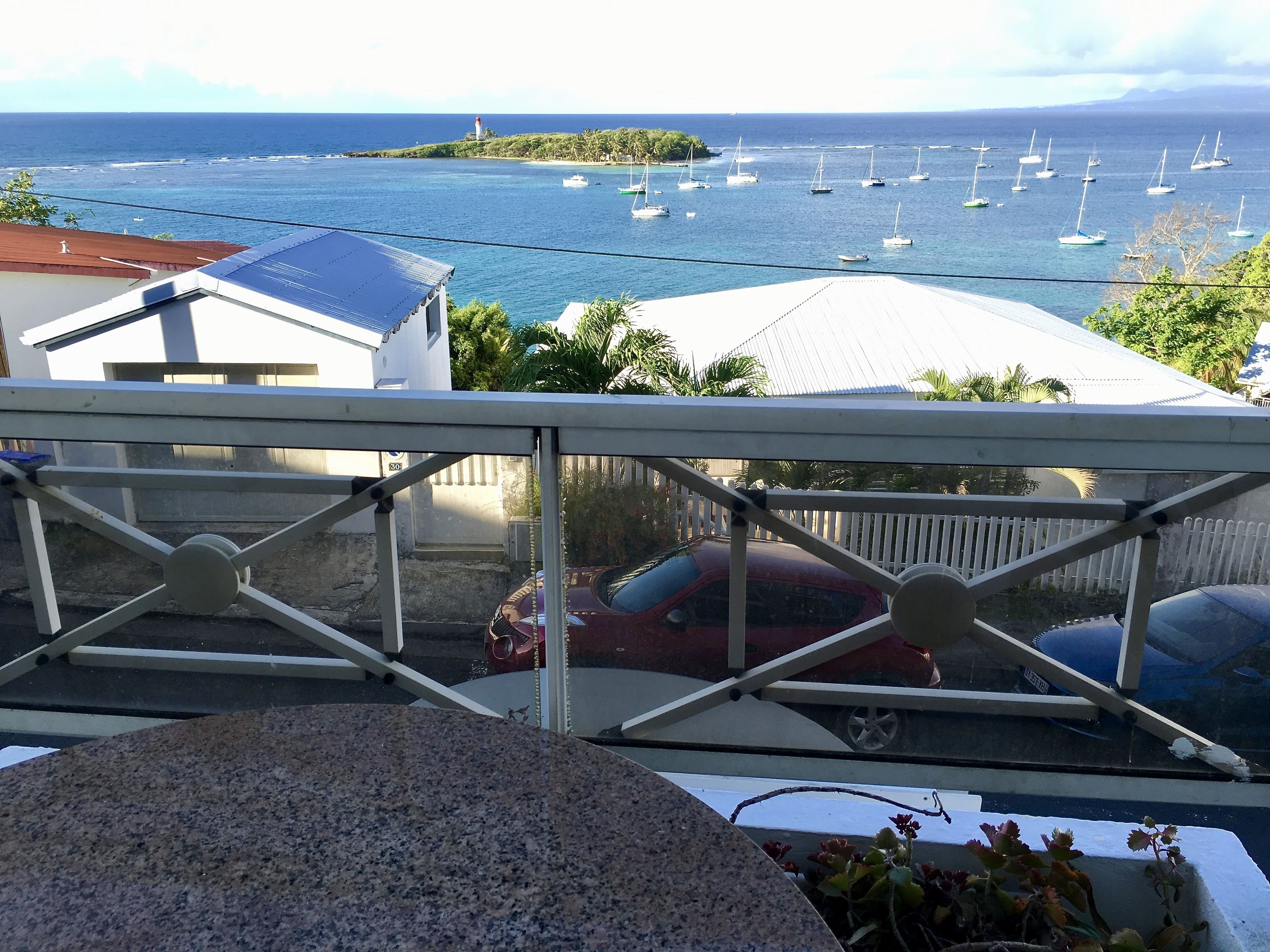 A balcony with a view of the sea, a red car, and a white fence.
