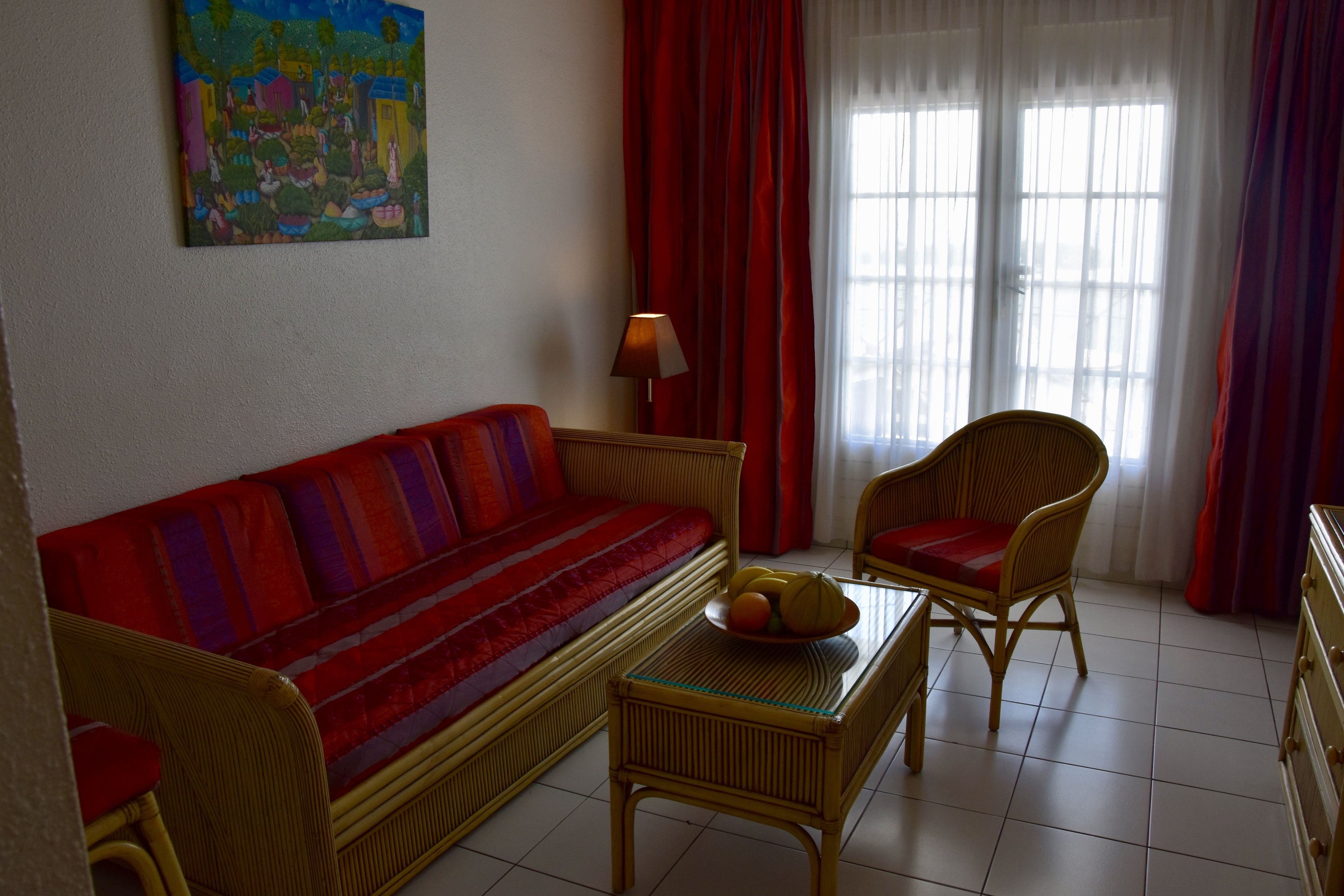 A living room with a red sofa, a wicker chair, a small table with a fruit bowl, and a painting on the wall.