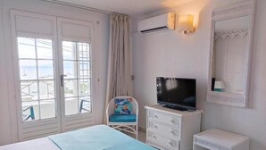 A hotel room with a bed, a television, a chair, a small table, and a view of the sea through large windows.