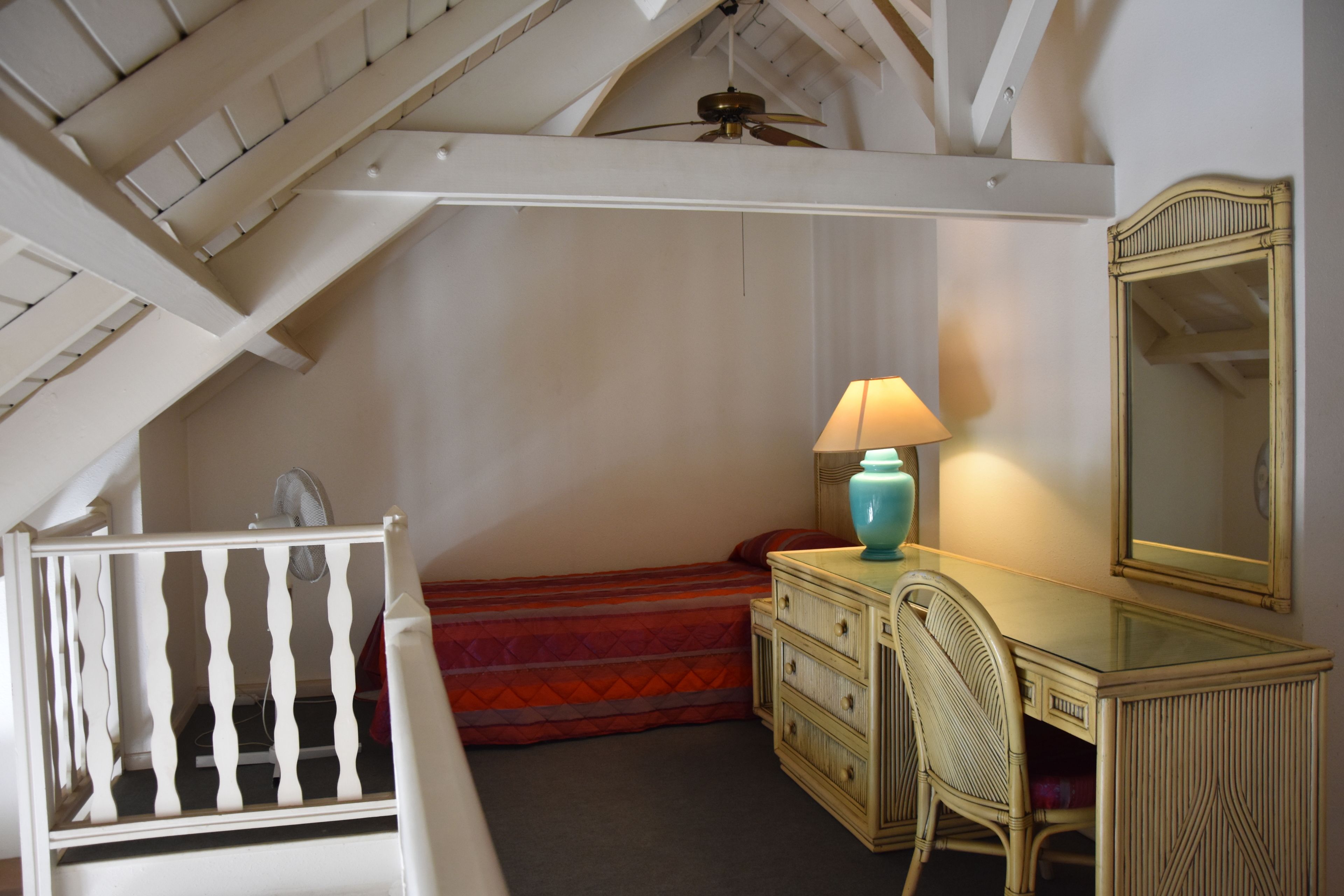 A bedroom with a sloped ceiling, a bed with a red bedspread, a dresser, a chair, a lamp, and a mirror.