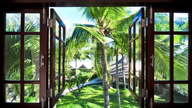 A view through a wooden-framed window with multiple panes, revealing a lush garden with palm trees and a thatched-roof structure. - Four Oceans Resort Muine (Mui Ne)