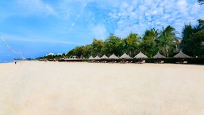 A beach with rows of thatched umbrellas and lounge chairs, palm trees, and a clear sky. - Four Oceans Resort Muine (Mui Ne)