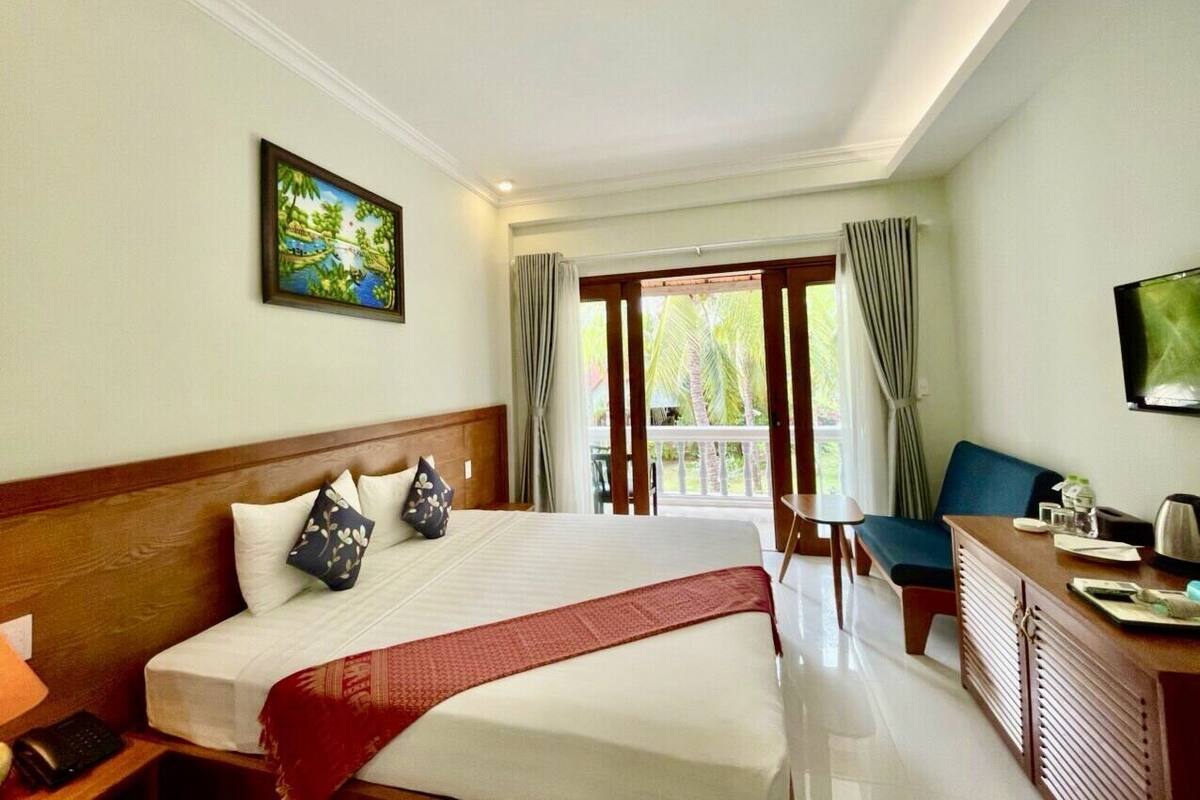 A hotel room with a large bed, a desk with a chair, a television, and a balcony with curtains.