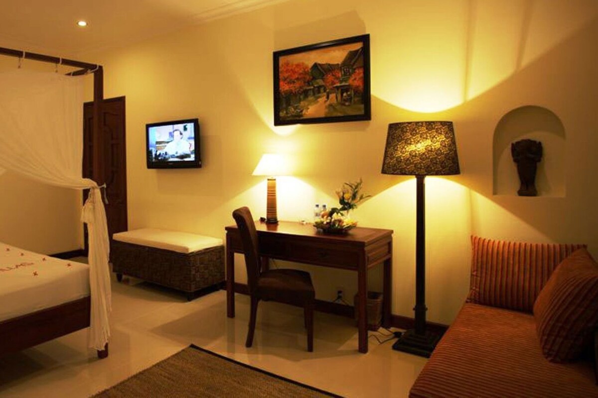 A hotel room with a four-poster bed, a desk with a chair, a sofa, a television, and a lamp.