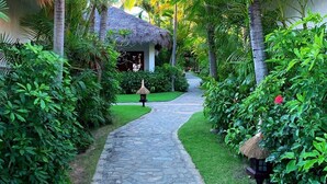 A stone pathway leading to a thatched-roof structure surrounded by lush greenery and palm trees. - Cham Villas (Mui Ne)