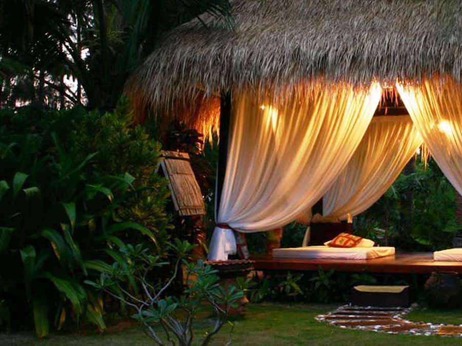 A thatched-roof outdoor bedroom with a bed, draped curtains, and ambient lighting.