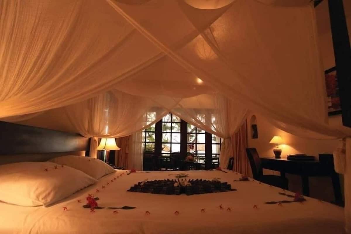 Quarto com cama coberta por lençóis brancos, arranjo circular de objetos sobre a cama e janela com cortinas.