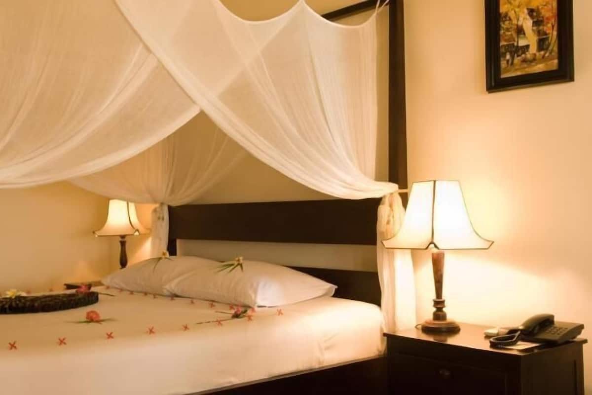 Uma cama com dossel, duas luminárias de cabeceira, um criado-mudo com telefone e um quadro na parede.
