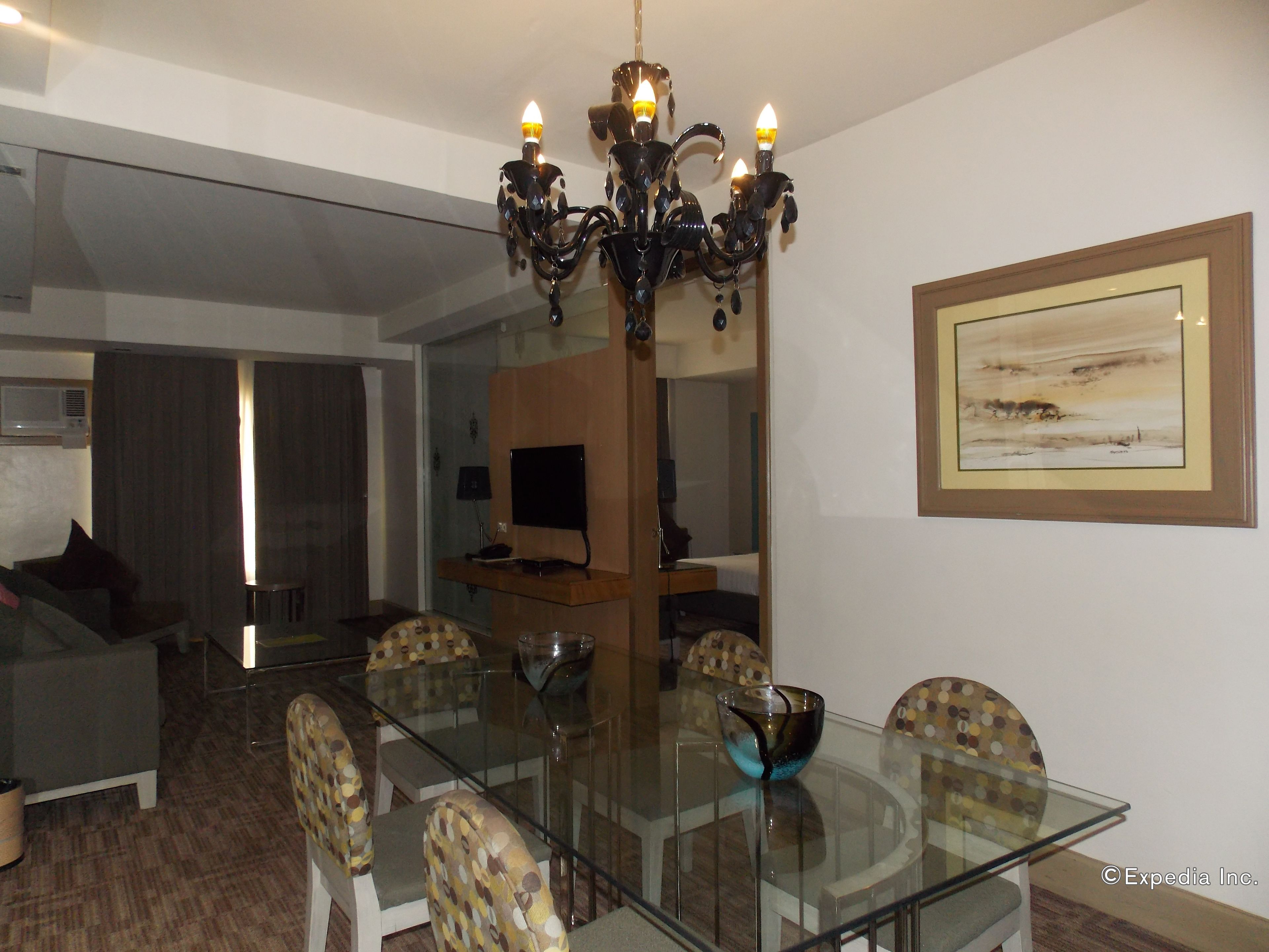 Comedor con mesa de vidrio, sillas, candelabro, televisor y un cuadro en la pared.