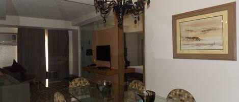 Comedor con mesa de vidrio, sillas, candelabro, televisor y un cuadro en la pared.