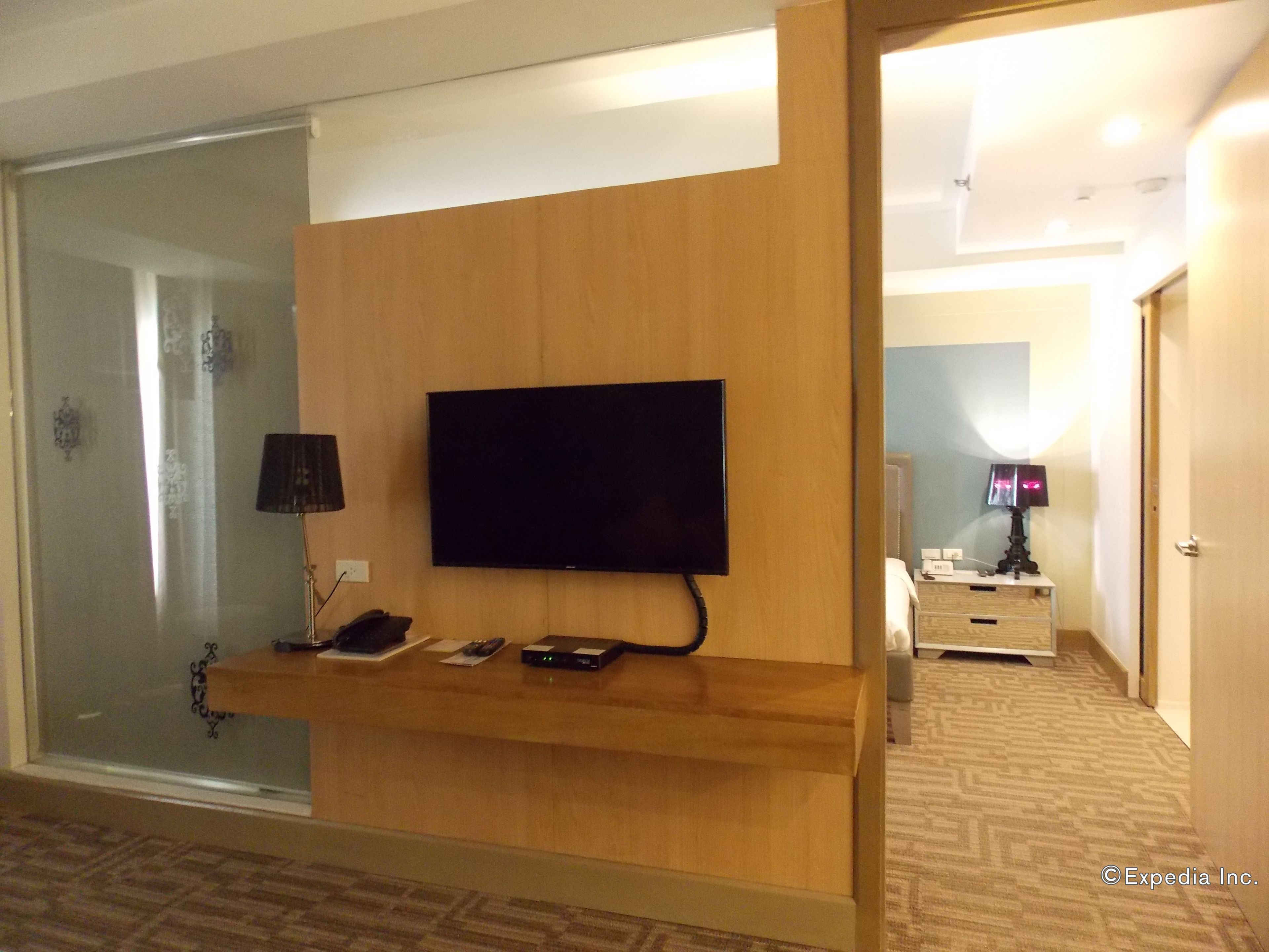 A hotel room with a wooden console table, a flat-screen TV, a lamp, and a glass partition.