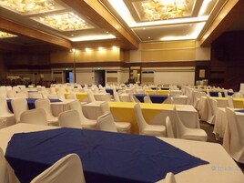 Un salón de conferencias con filas de mesas y sillas, equipado con luces en el techo y una pantalla para proyector.
