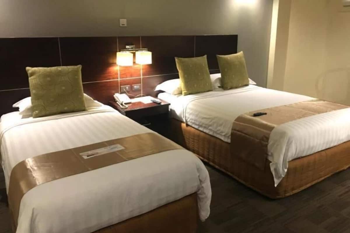 A hotel room with two beds, a nightstand, a lamp, and a wall-mounted light.