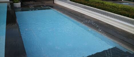 Une piscine rectangulaire à l’eau claire, entourée d’une allée pavée et d’une haie basse.