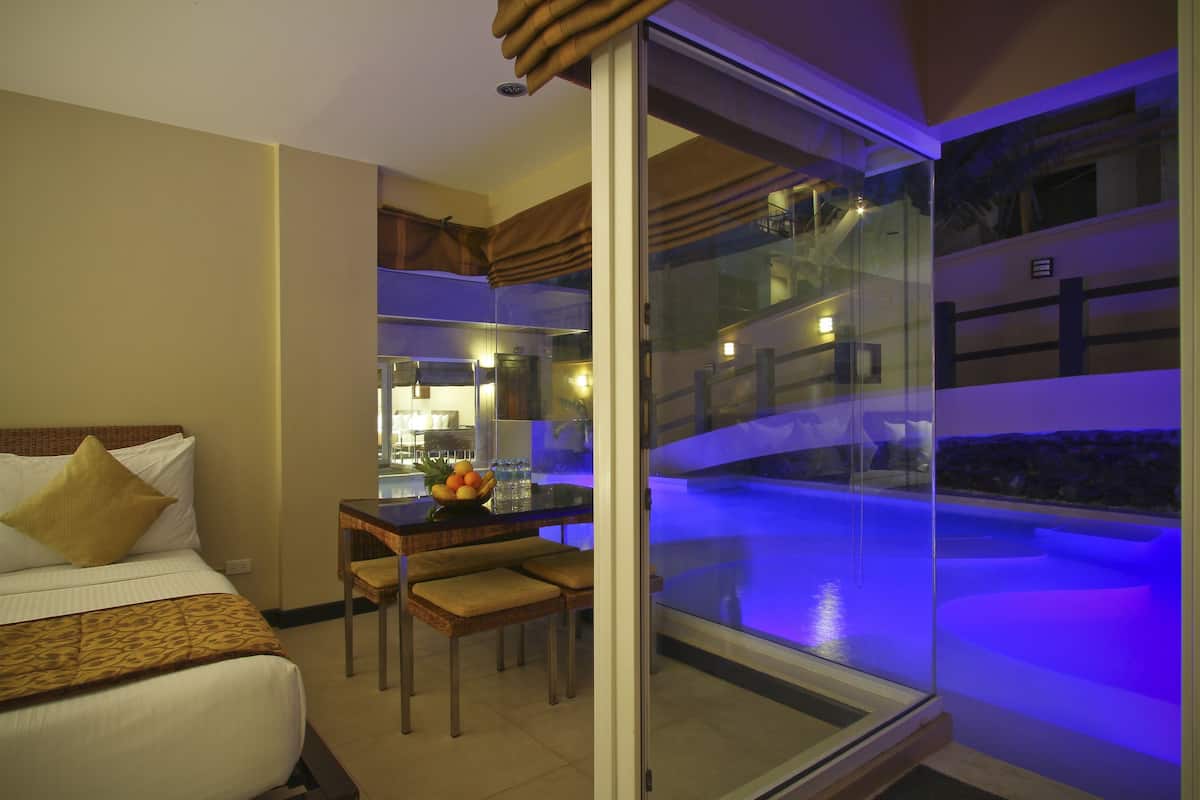 Habitación de hotel con cama, una mesa de comedor con sillas, una pequeña cocina y un balcón con vidrio que da a una piscina.