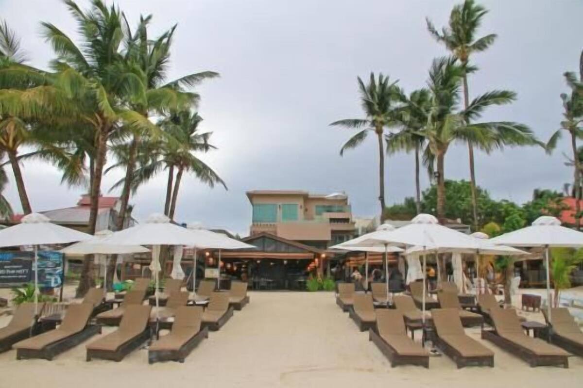 Un resort frente a la playa con sillones y sombrillas, palmeras y un edificio al fondo.