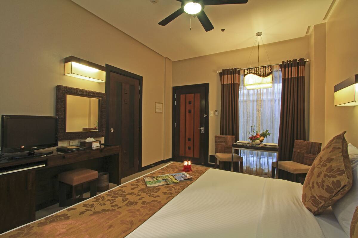 Habitación de hotel con cama, escritorio con televisor, lámpara de escritorio, silla, mesa con un jarrón de flores y ventilador de techo.