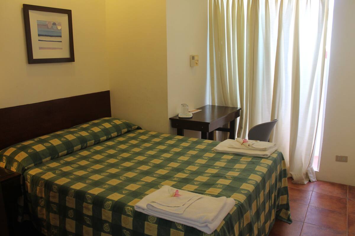 Une chambre d’hôtel avec une couvre-lit à carreaux, un bureau avec une chaise, un tableau encadré au mur et une fenêtre avec des rideaux.