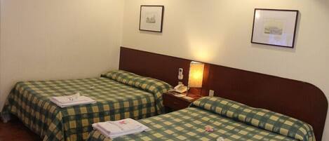 A hotel room with two single beds, a bedside table with a lamp, a wall-mounted air conditioner, and framed pictures on the walls.