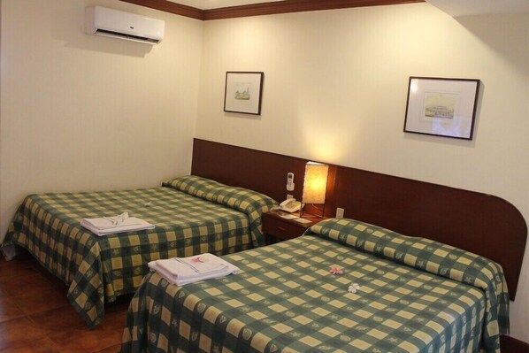 A hotel room with two single beds, a bedside table with a lamp, a wall-mounted air conditioner, and framed pictures on the walls.