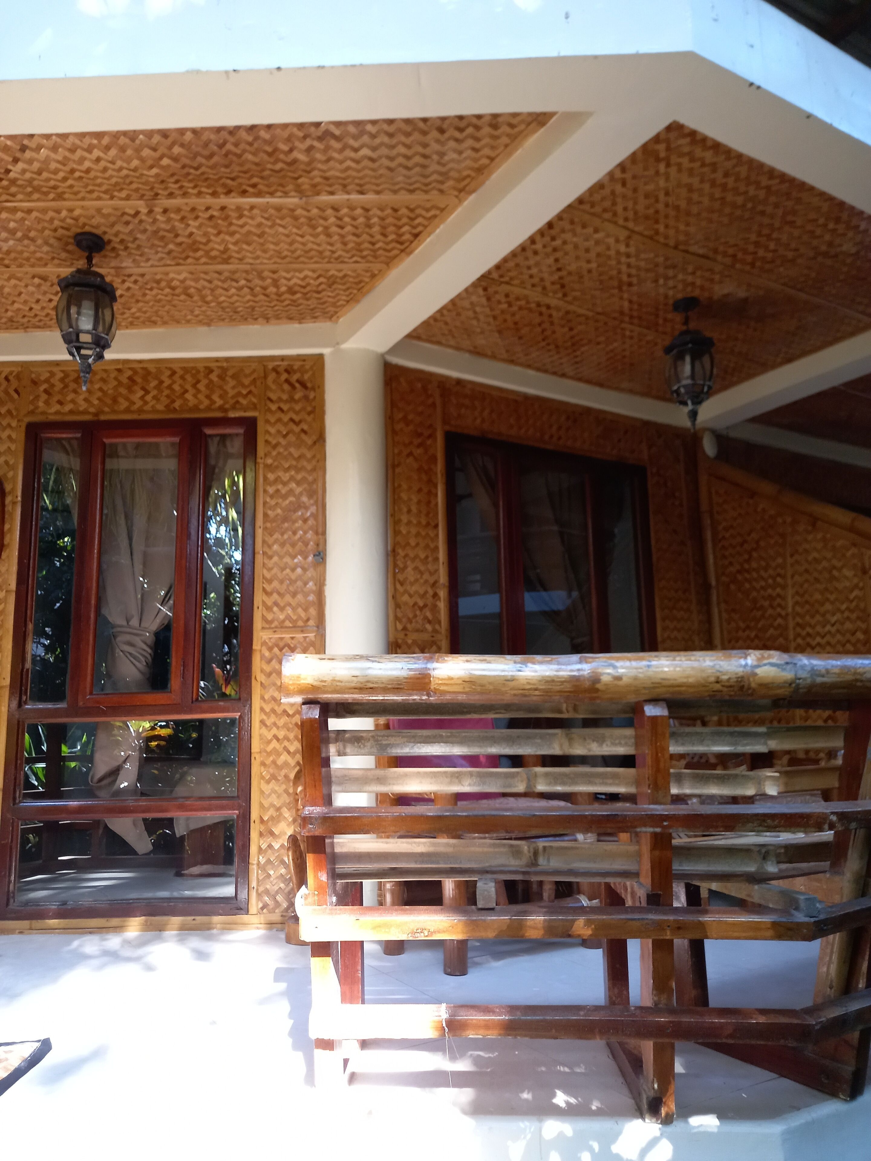 A wooden bench outside a building with a textured wall and two windows with curtains.