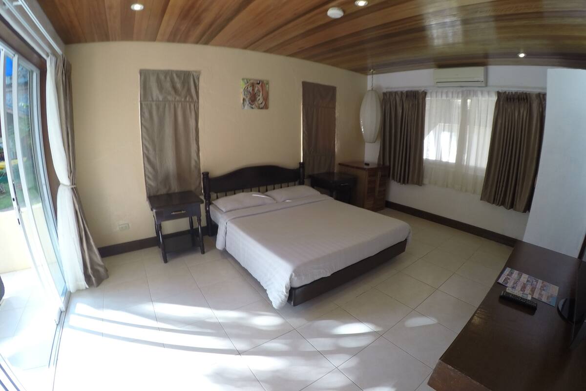 Un dormitorio con una cama grande, un escritorio, una silla y un armario. Hay una ventana con cortinas, un cuadro en la pared y un equipo de aire acondicionado.