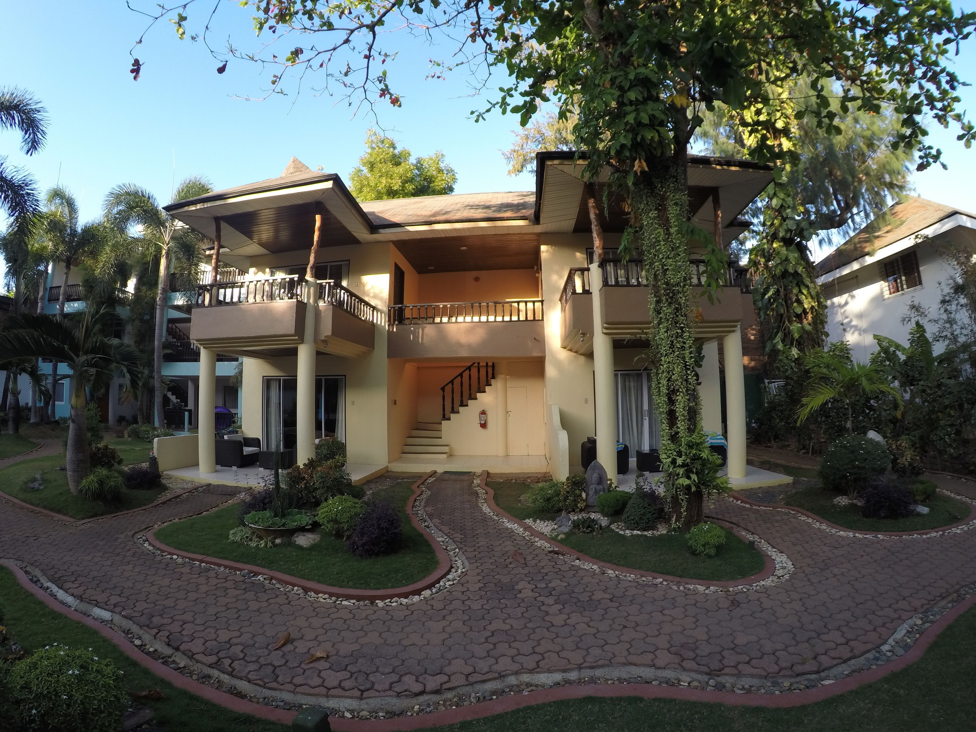 A two-story house with a central staircase, surrounded by a garden with palm trees and shrubs.