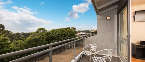 A balcony with a glass railing, two white chairs, and a view of trees and buildings.