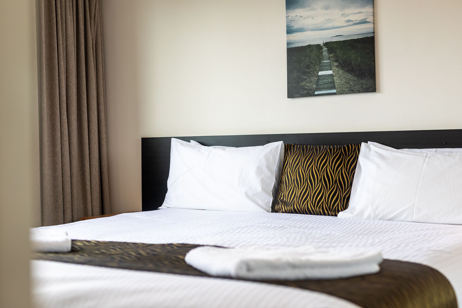 A neatly made bed with white bedding and a patterned pillow, a wooden headboard, and a painting of a beach scene on the wall.