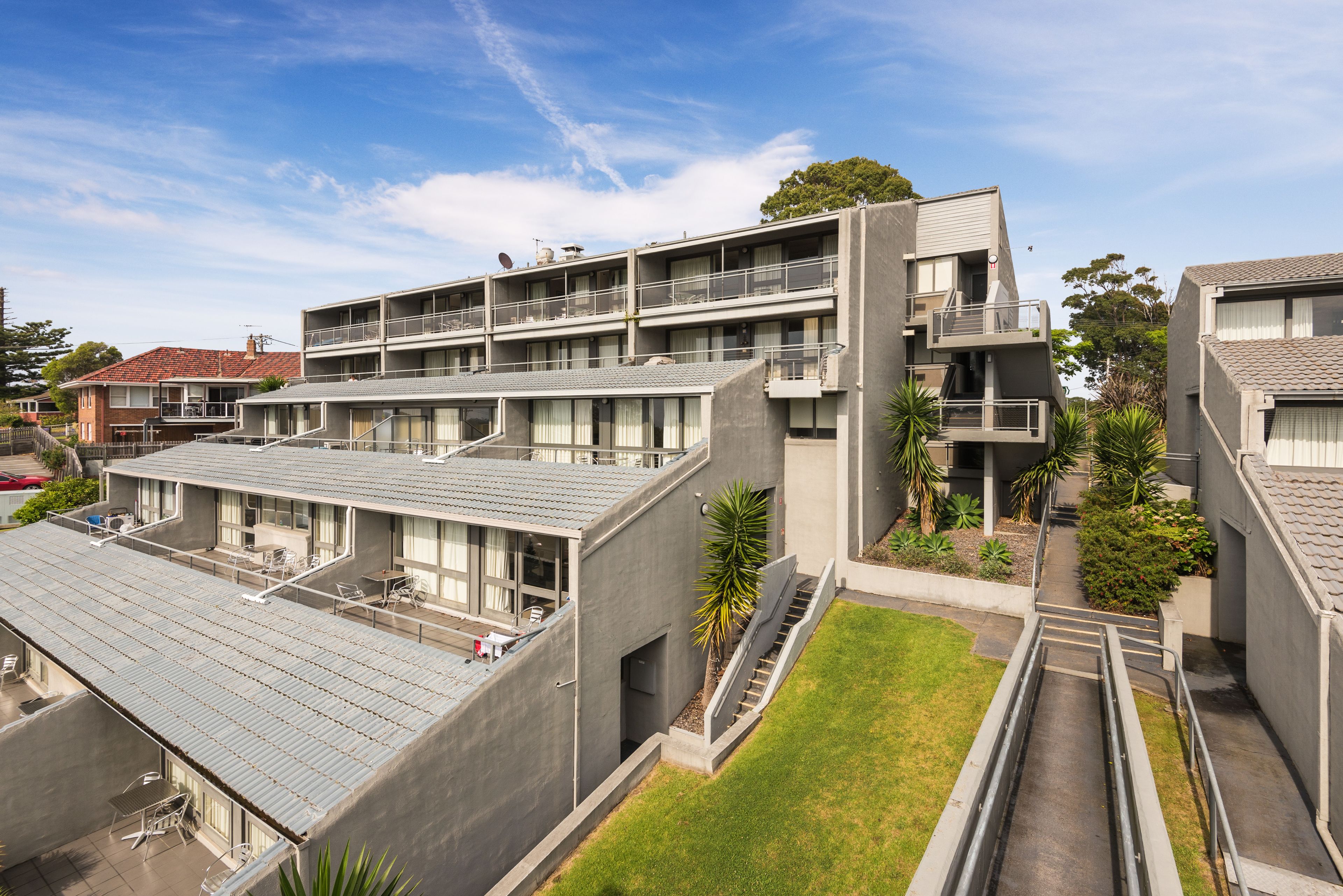 A modern multi-story building with a flat roof, balconies, and a small garden area.