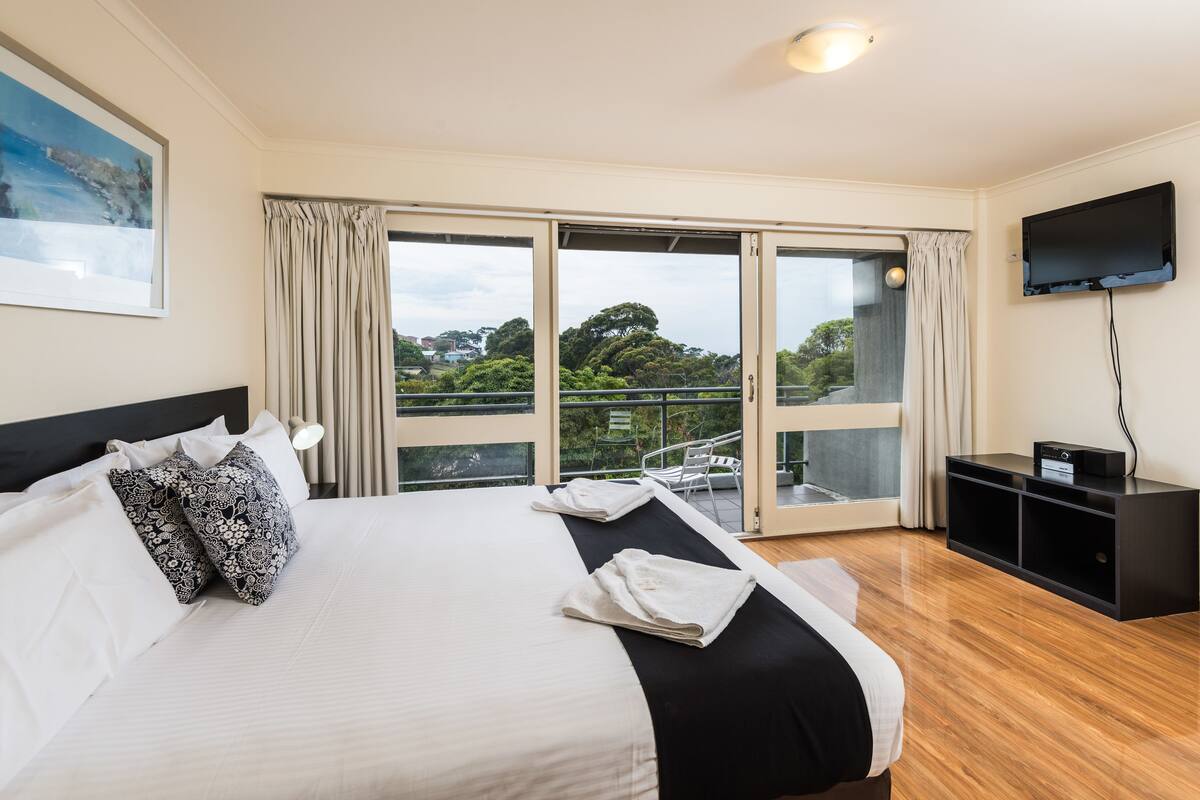 A hotel room with a large bed, a flat-screen TV, a balcony with a view of trees, and a sliding glass door.