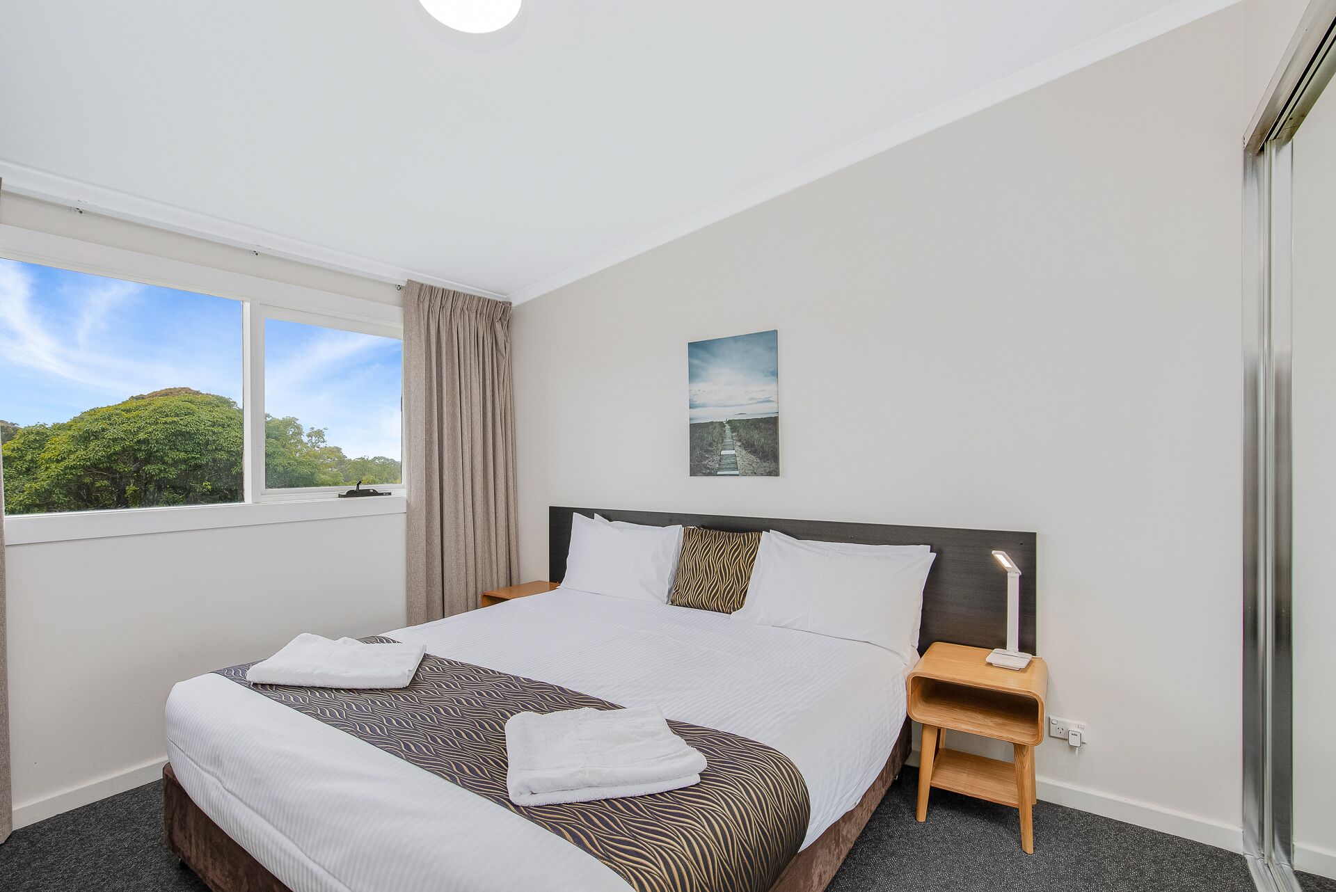 A hotel room with a large bed, a bedside table, a nightstand, a window with a view of trees, and a painting on the wall.