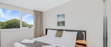 A hotel room with a large bed, a bedside table, a nightstand, a window with a view of trees, and a painting on the wall.