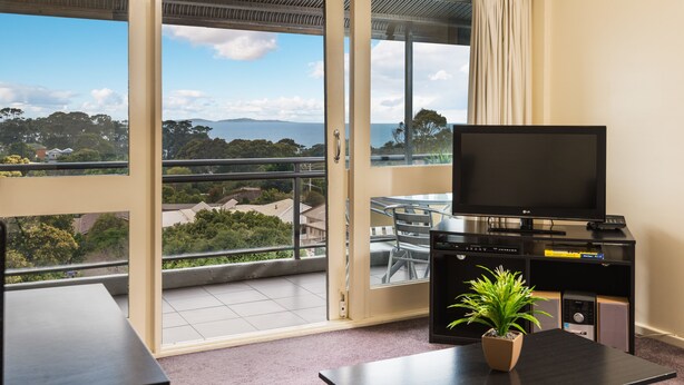 A balcony with a view of the sea and hills, a TV on a stand, a table, and a potted plant.