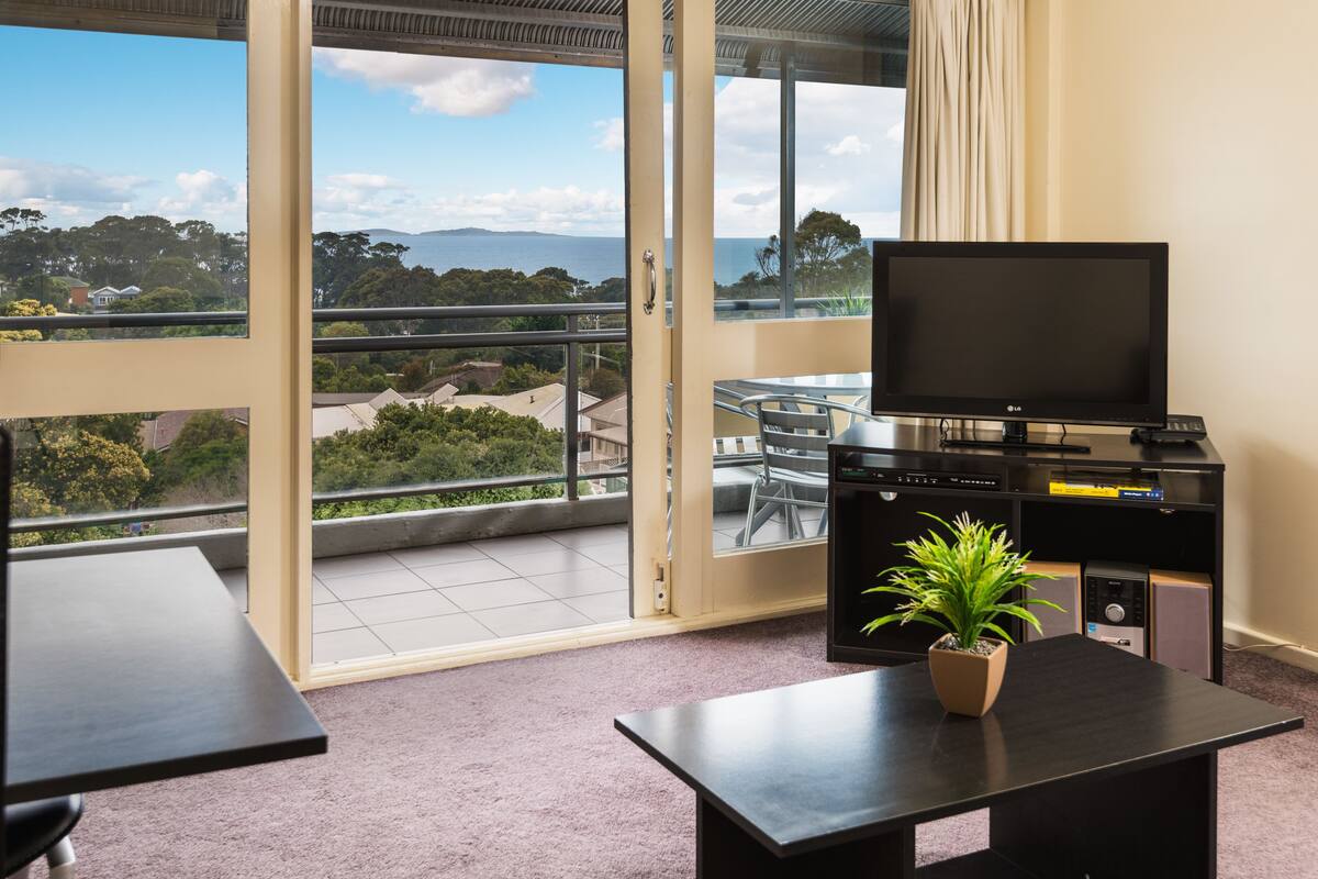 A balcony with a view of the sea and hills, a TV on a stand, a table, and a potted plant.