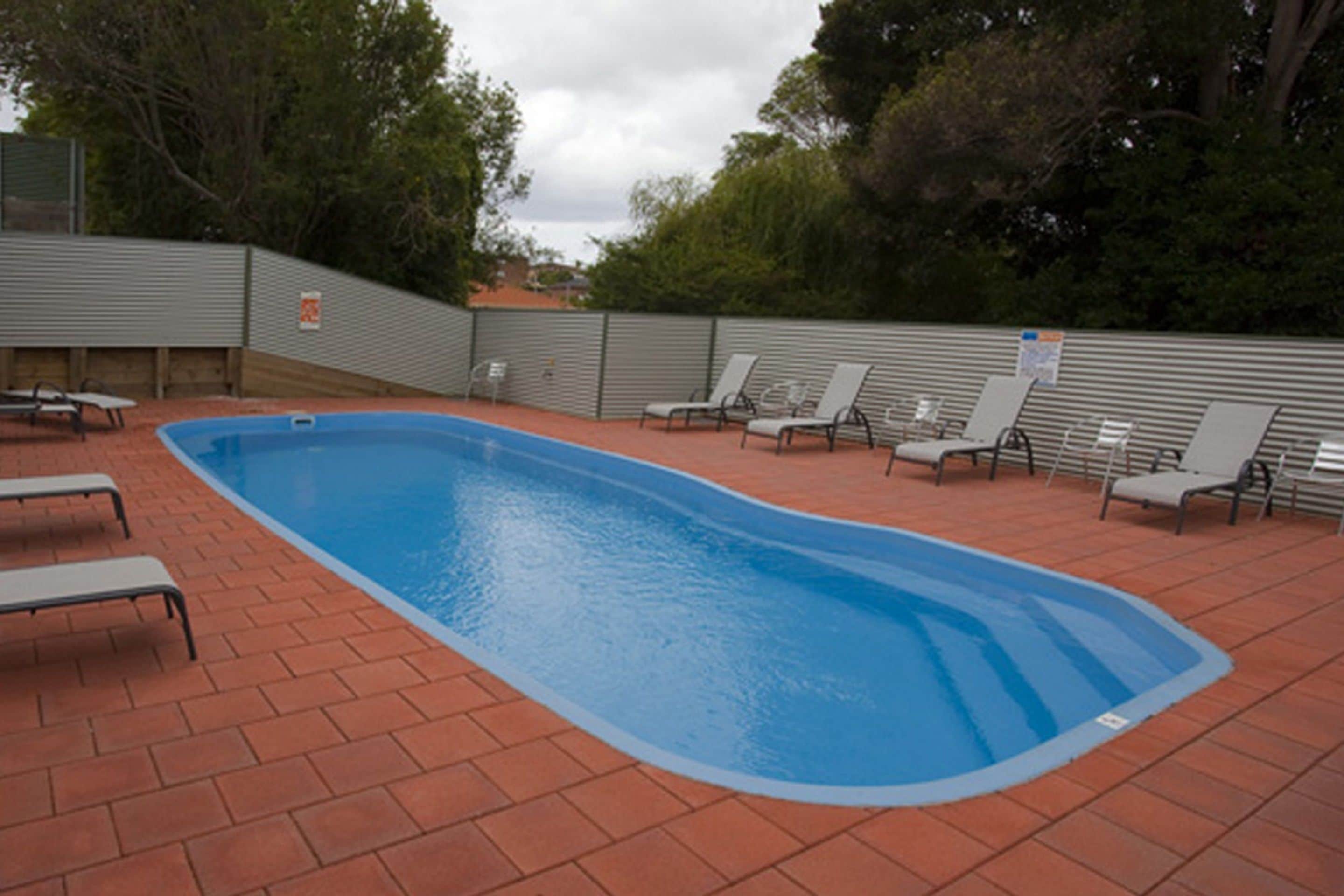 Une piscine rectangulaire aux parois bleues, entourée d’une terrasse en briques. On y trouve plusieurs chaises longues et une table de pique-nique.