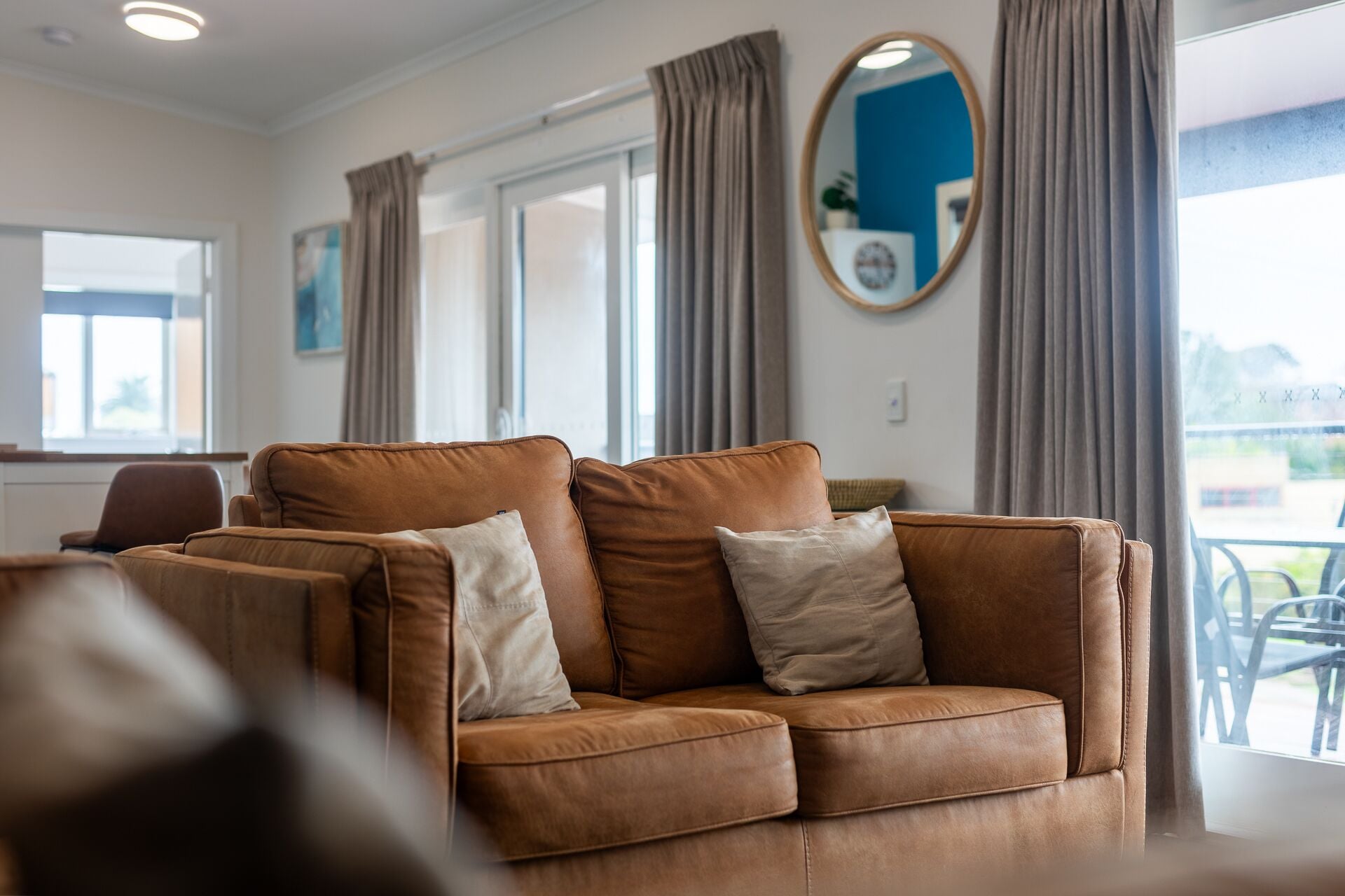 Een bruine leren bank met kussens in een heldere kamer met grote ramen en gordijnen.