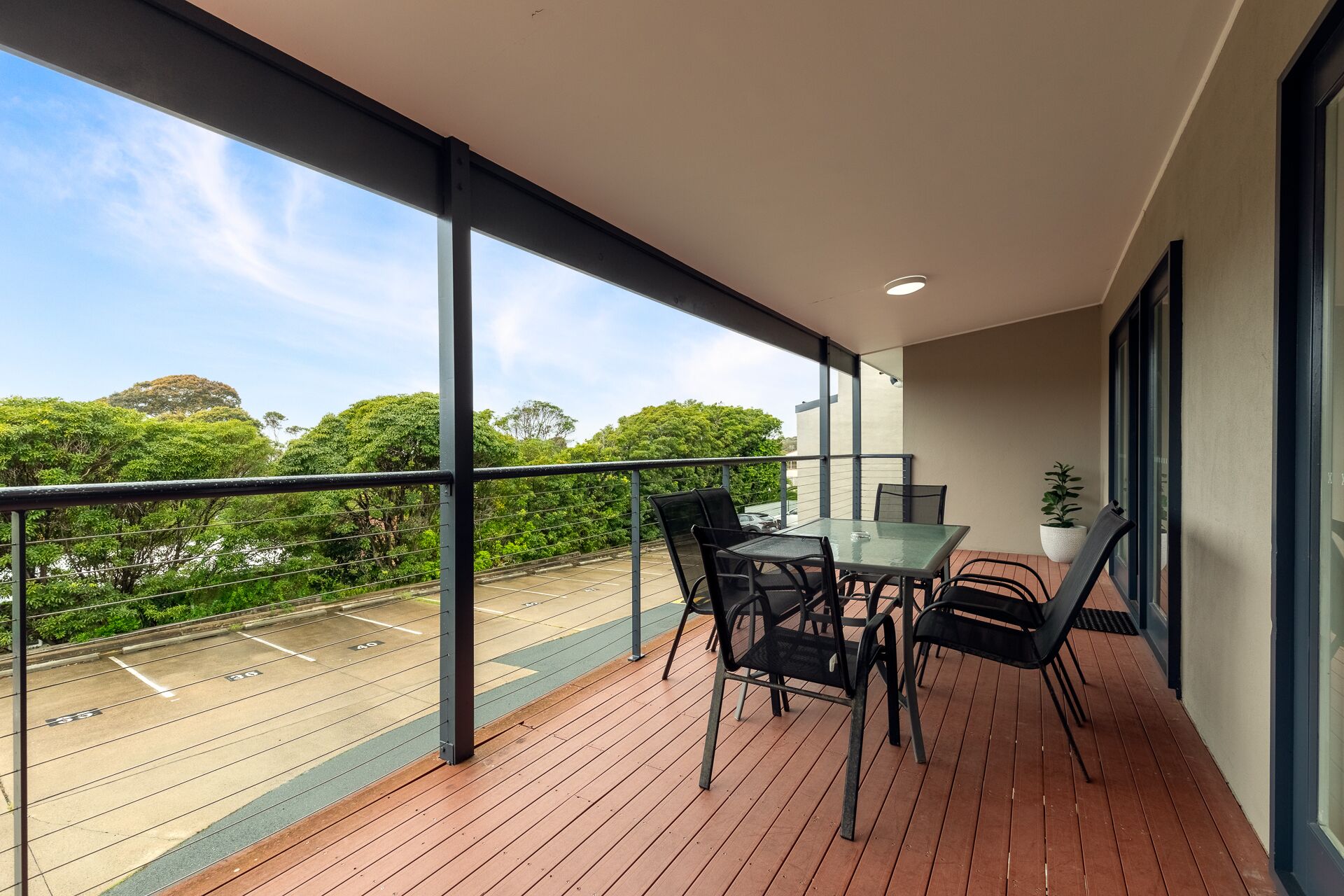 A balcony with a table and chairs, a potted plant, and a view of a parking area and trees.