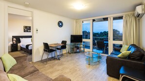 A modern living room with a sofa, a dining table, and a TV. There is a balcony with a view of trees.