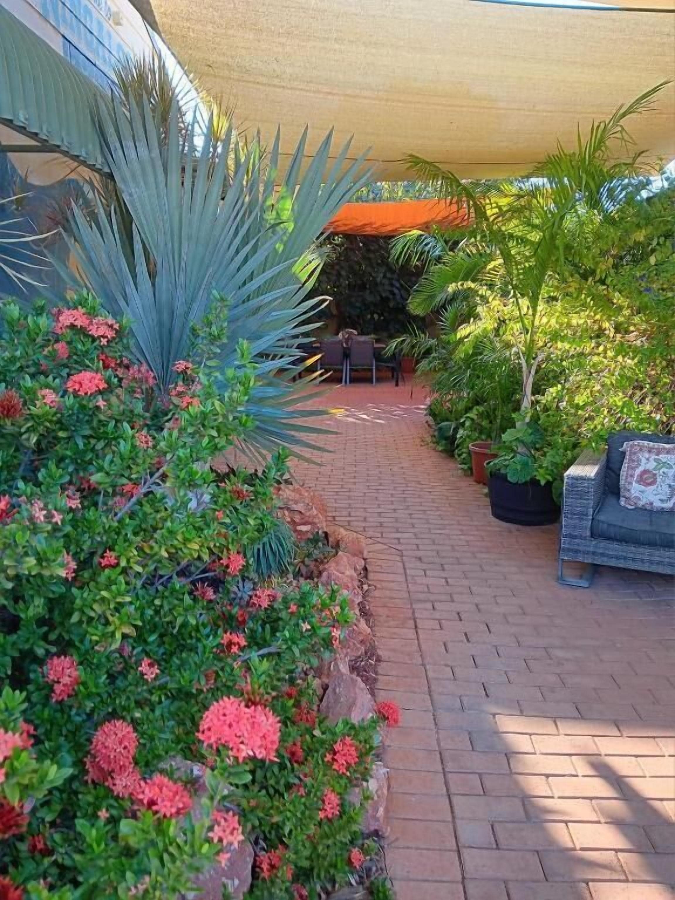 A patio area with a brick pathway, potted plants, and a seating area under a shade structure.