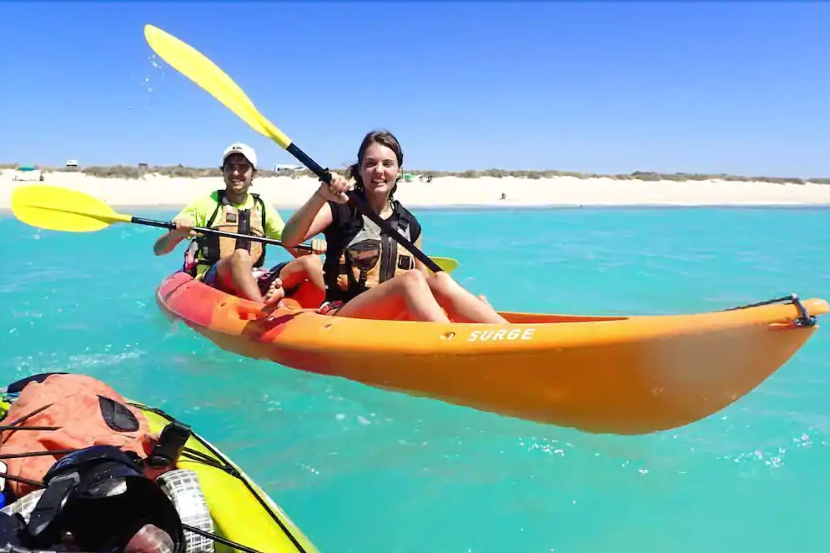 Two people kayaking on a clear blue sea with a sandy beach in the background.