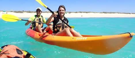 Deux personnes pratiquent le kayak sur une mer d’un bleu limpide, avec une plage de sable en arrière-plan.