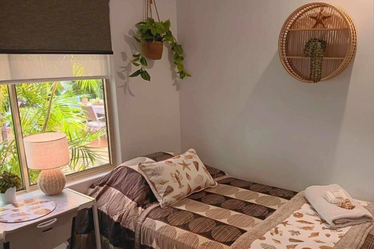 A bedroom with a bed, a desk, a lamp, a hanging plant, and a wall-mounted shelf.