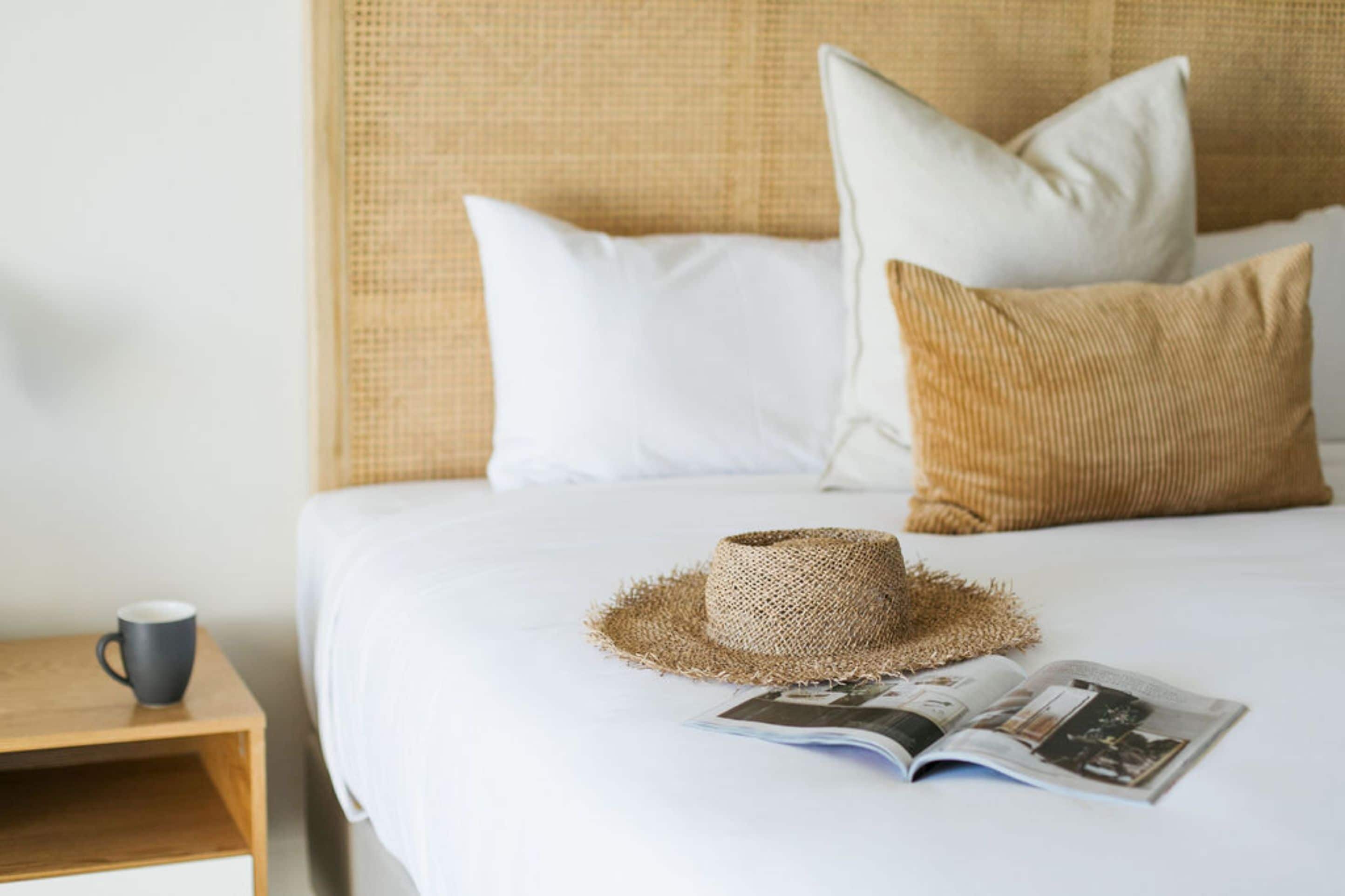 A neatly made bed with white bedding, a straw hat, and a magazine on the bed.