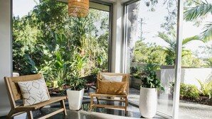 Un interior modern amb dues cadires de fusta i una banca blanca, envoltat de grans finestres i plantes en testos.